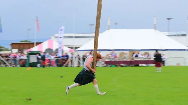 A competitor in a kilt running with a long wooden caber on the grass at a Highland gathering