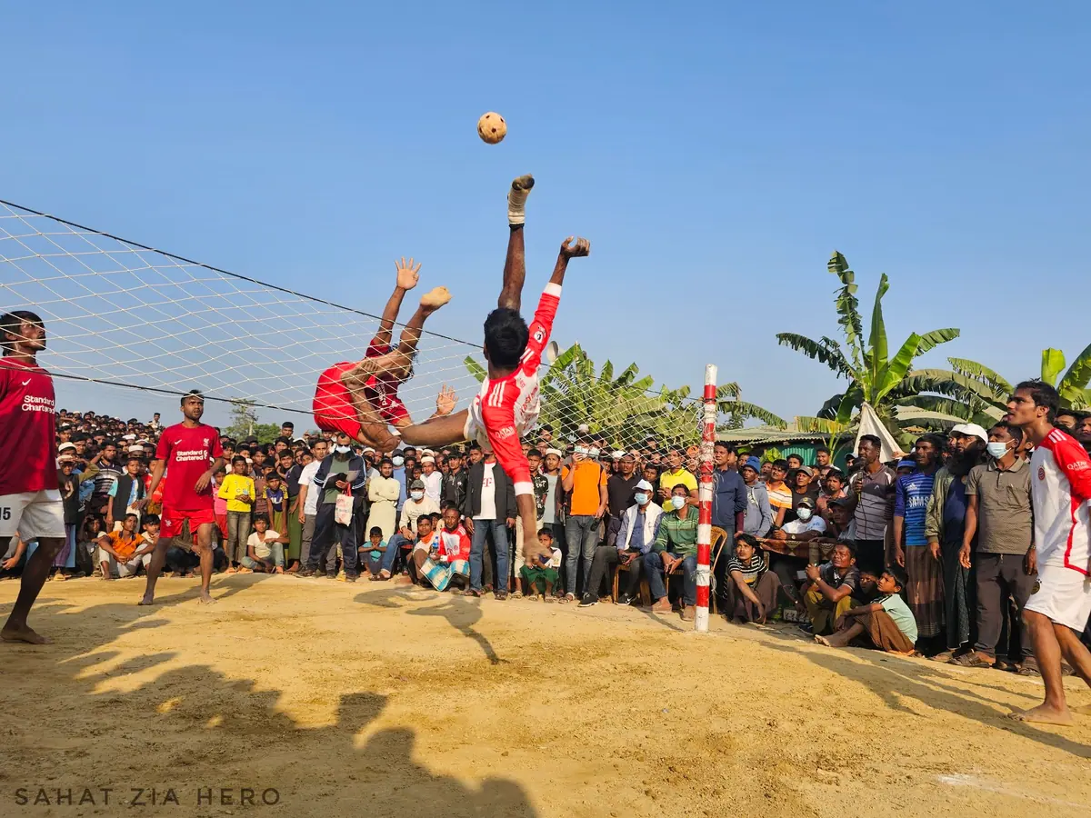 Chinlone players in a circle kicking a woven ball over the net, with a large crowd watching