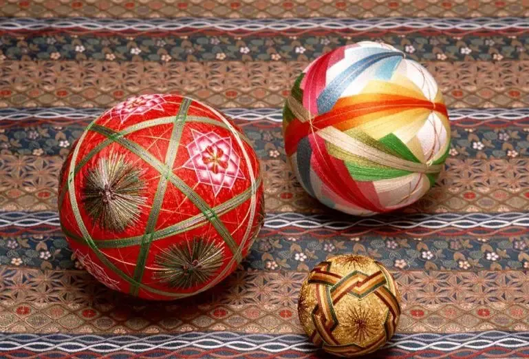 Three decorated cuju balls in red, multicolour and gold, with tasselled seams, resting on patterned cloth