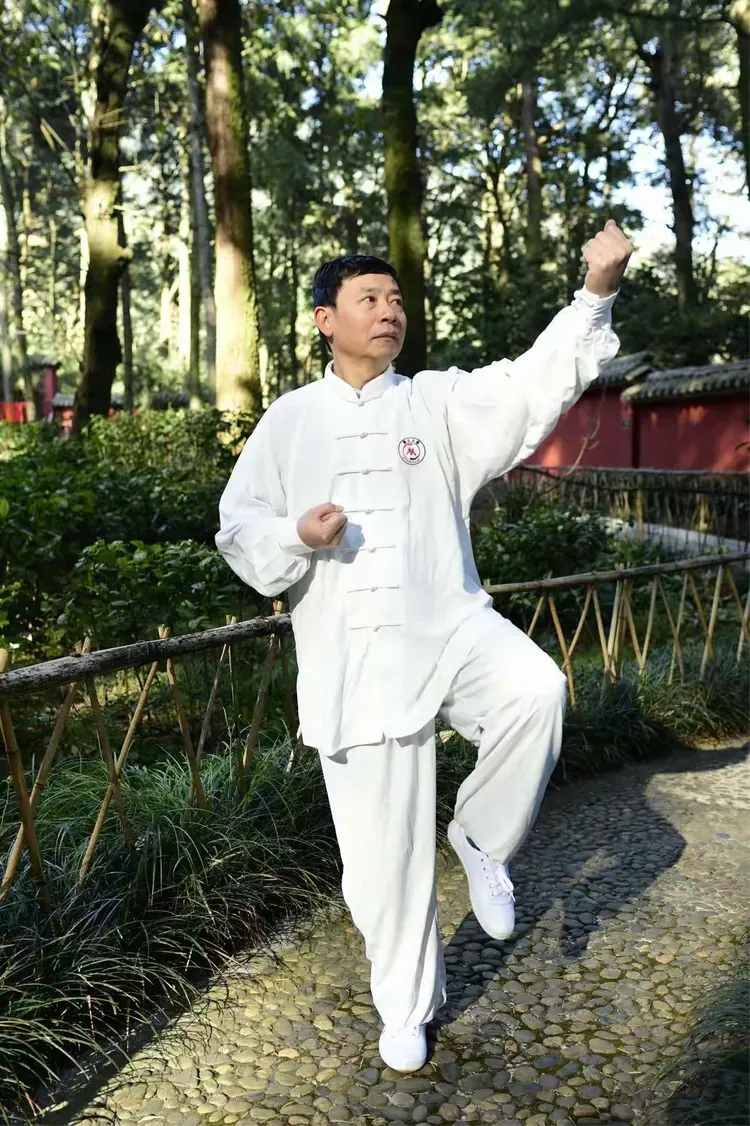 A practitioner in a white Tai Chi uniform with one knee raised and one fist lifted, practising in a garden